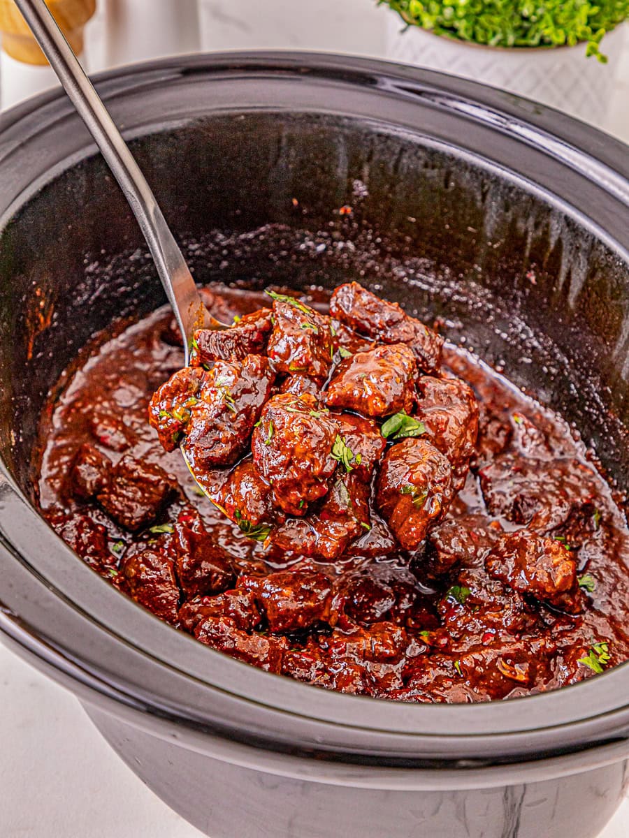 A close-up of beef stew with sauce and herbs in a black slow cooker, with a metal ladle lifting a portion.