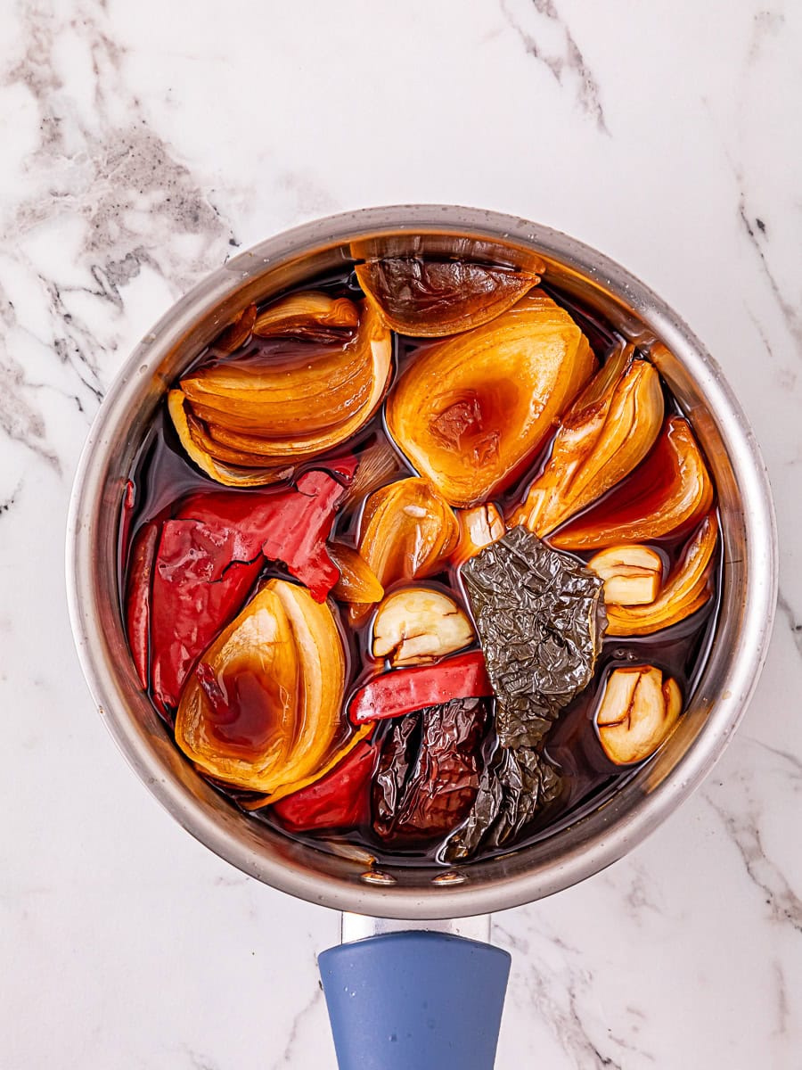 A saucepan filled with onion, garlic, dried and fresh chili peppers soaking in a dark liquid on a marble countertop.