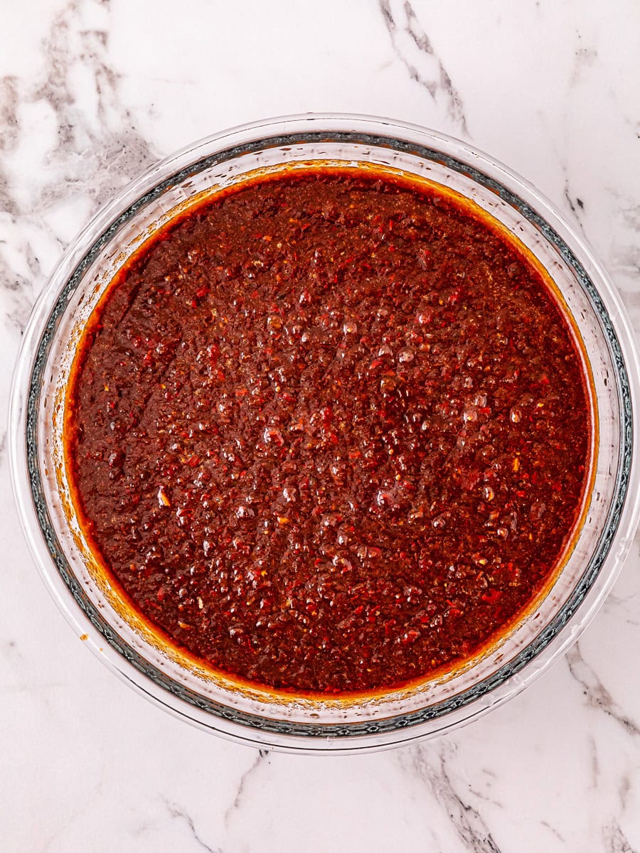 A clear glass bowl filled with a thick, reddish-brown chili paste sits on a white marble surface.
