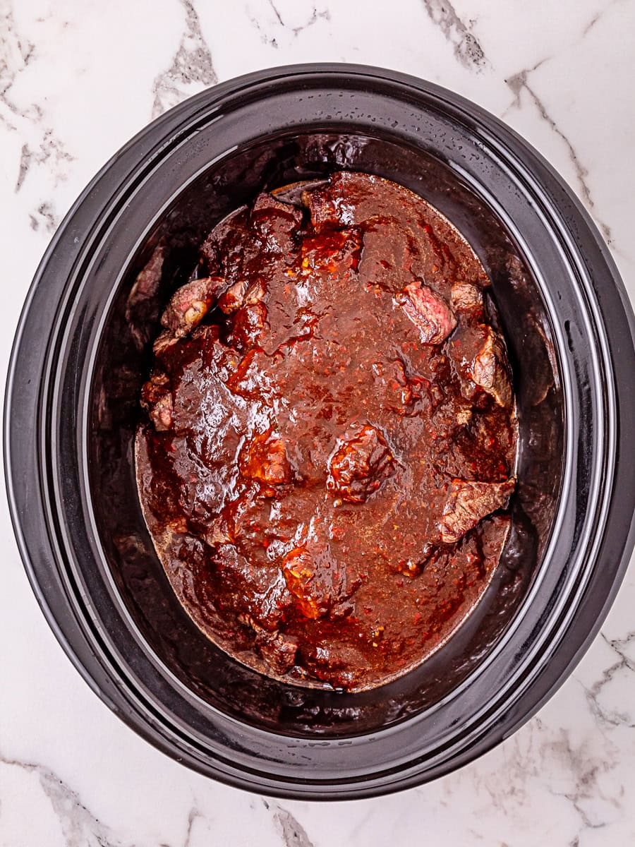 Slow cooker filled with chunks of beef in a thick, dark red sauce, placed on a white marble surface.