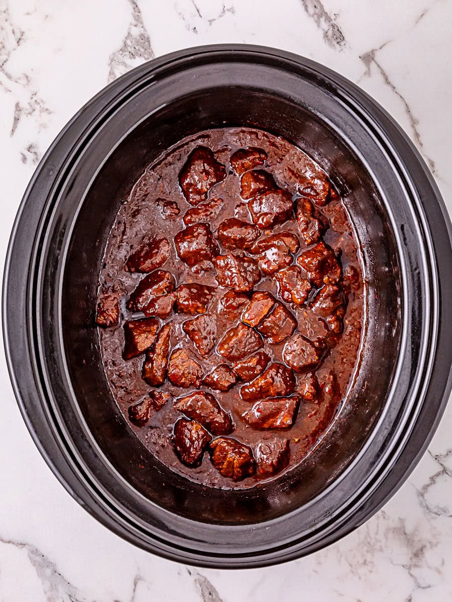 Chunks of beef in a dark sauce are cooked in a black slow cooker, shown from above on a white marble surface.