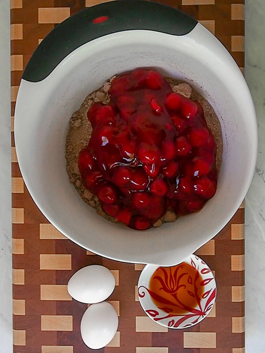 A mixing bowl with chocolate cake mix and cherry pie filling, next to two eggs and a small bowl of vanilla extract on a checkered cutting board.