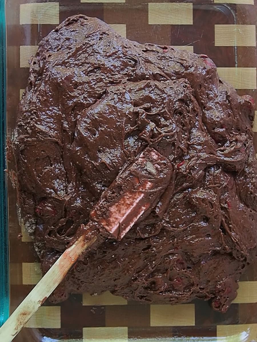Thick chocolate batter with visible chunks being spread in a glass baking dish using a spatula.