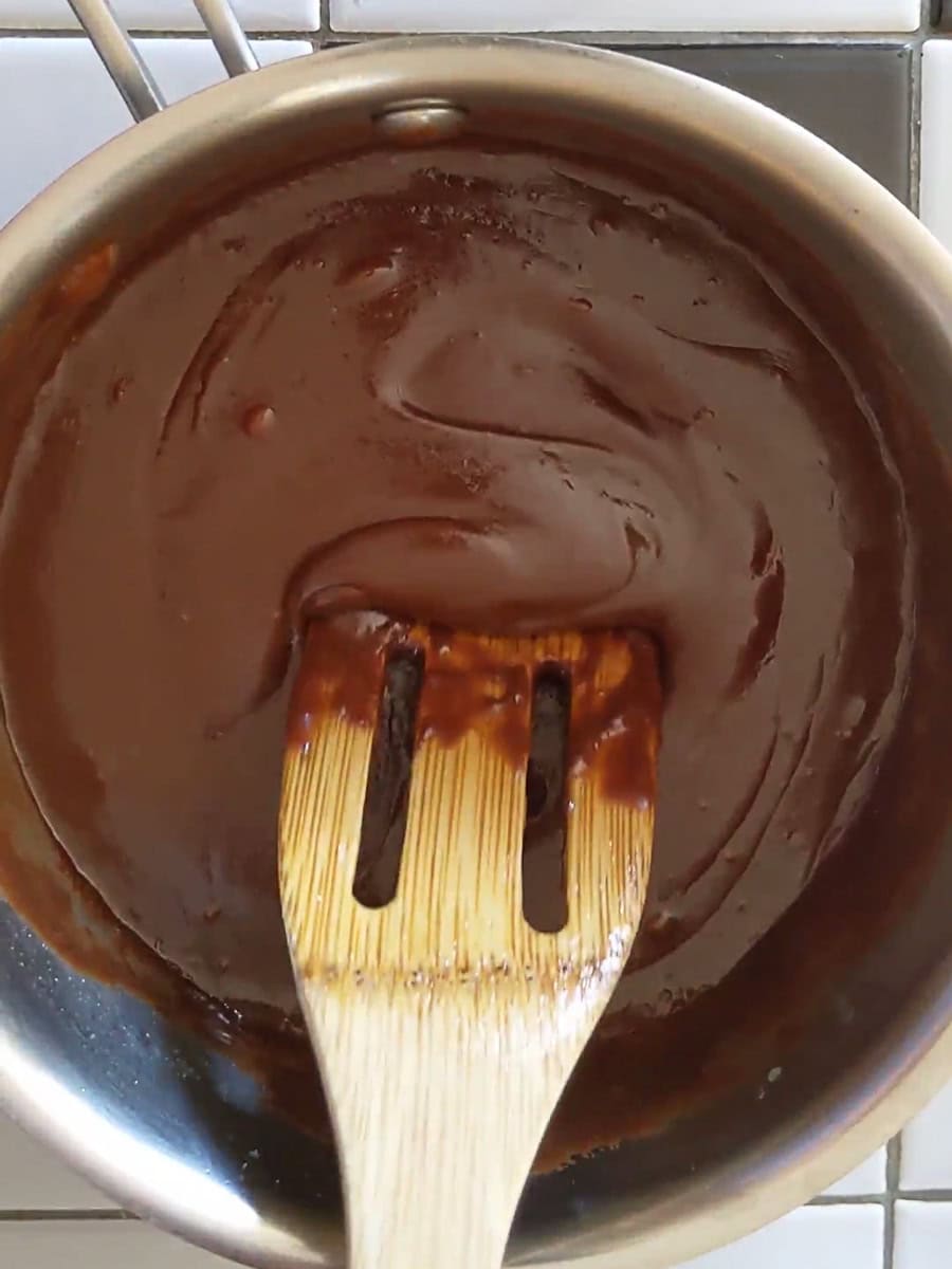 A wooden spatula stirs thick, smooth chocolate sauce in a stainless steel saucepan on a tiled countertop.