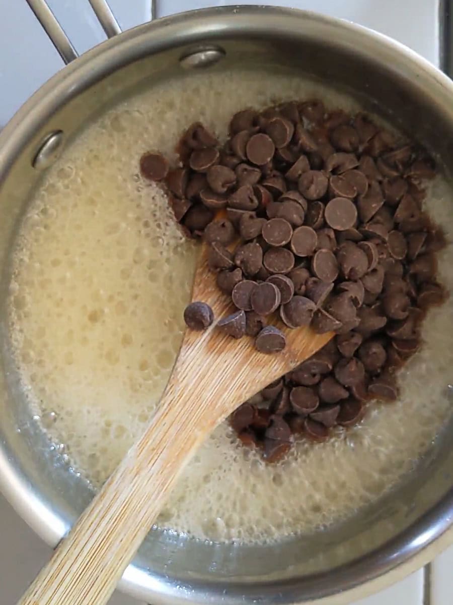 Chocolate chips are being stirred into a boiling mixture in a saucepan with a wooden spoon.