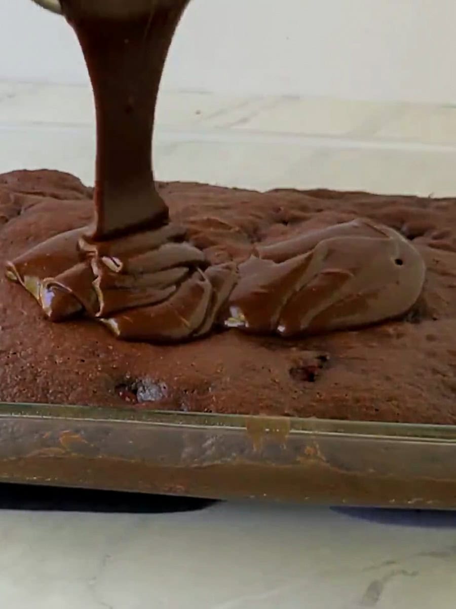 Chocolate frosting is being poured onto a rectangular chocolate cake in a glass baking dish.