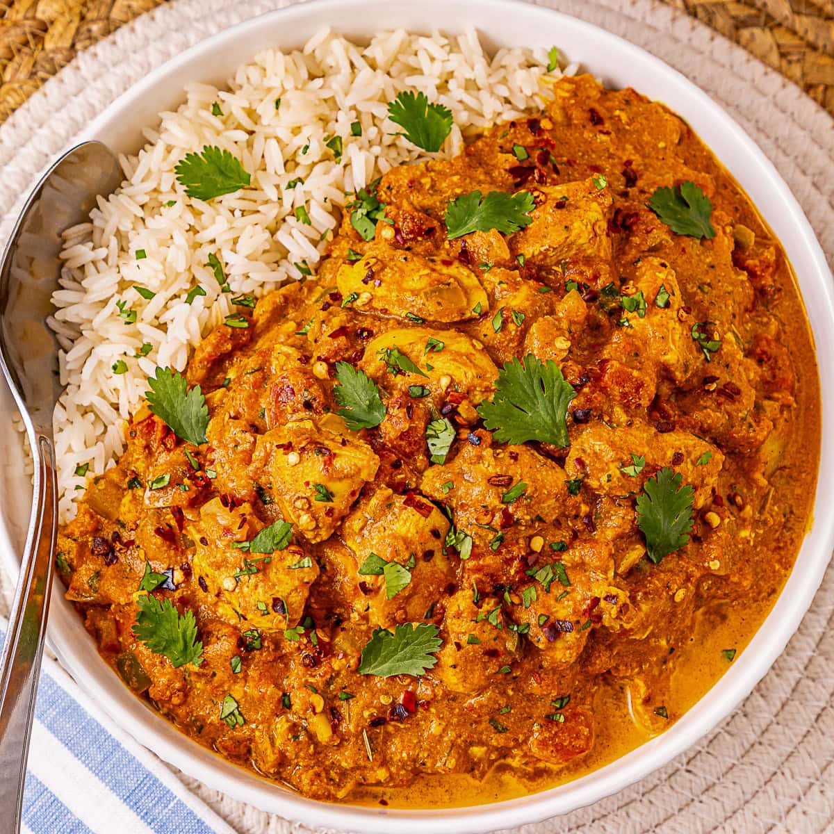 A bowl of white rice and creamy chicken curry garnished with cilantro, served with a spoon.