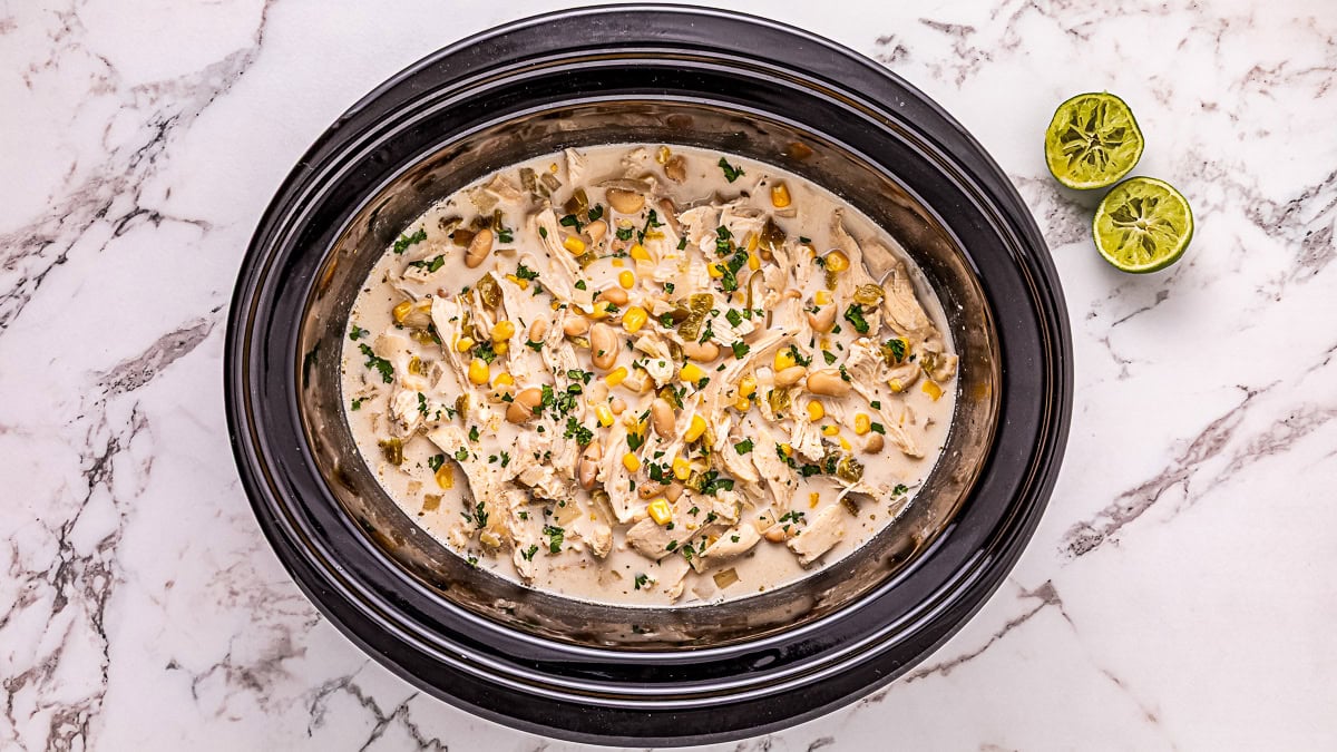 A slow cooker filled with creamy shredded chicken, corn, and beans, garnished with herbs, sits on a marble countertop next to two squeezed lime halves.