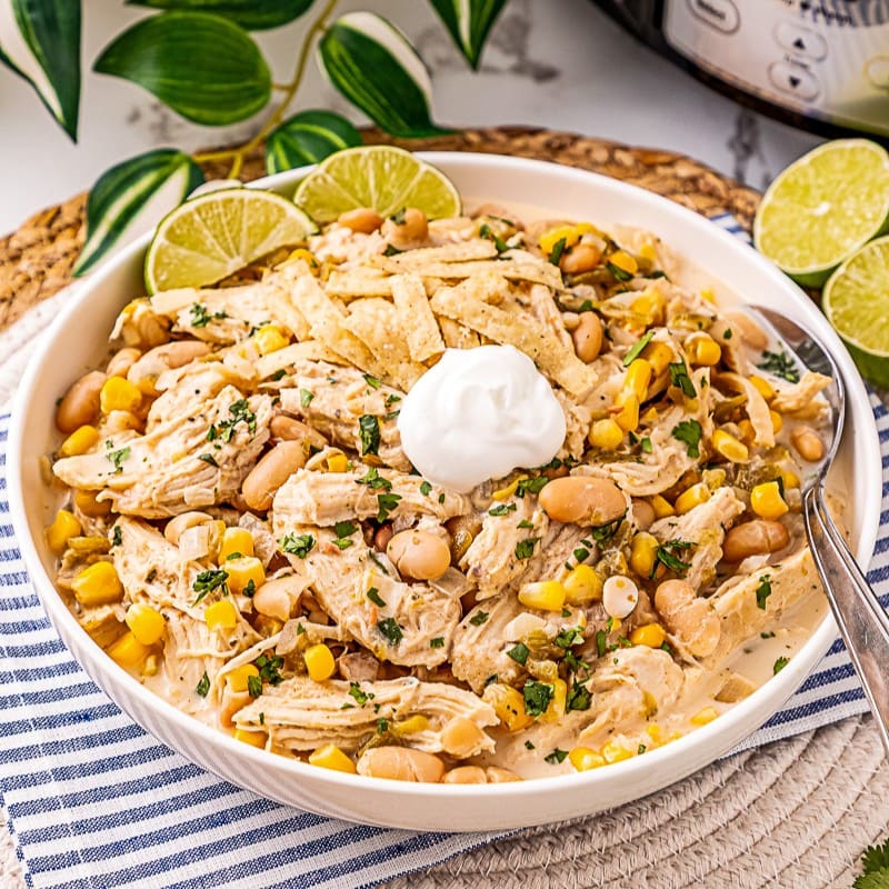 A bowl of creamy chicken chili with shredded chicken, corn, white beans, tortilla strips, and a dollop of sour cream, garnished with chopped herbs and lime slices.