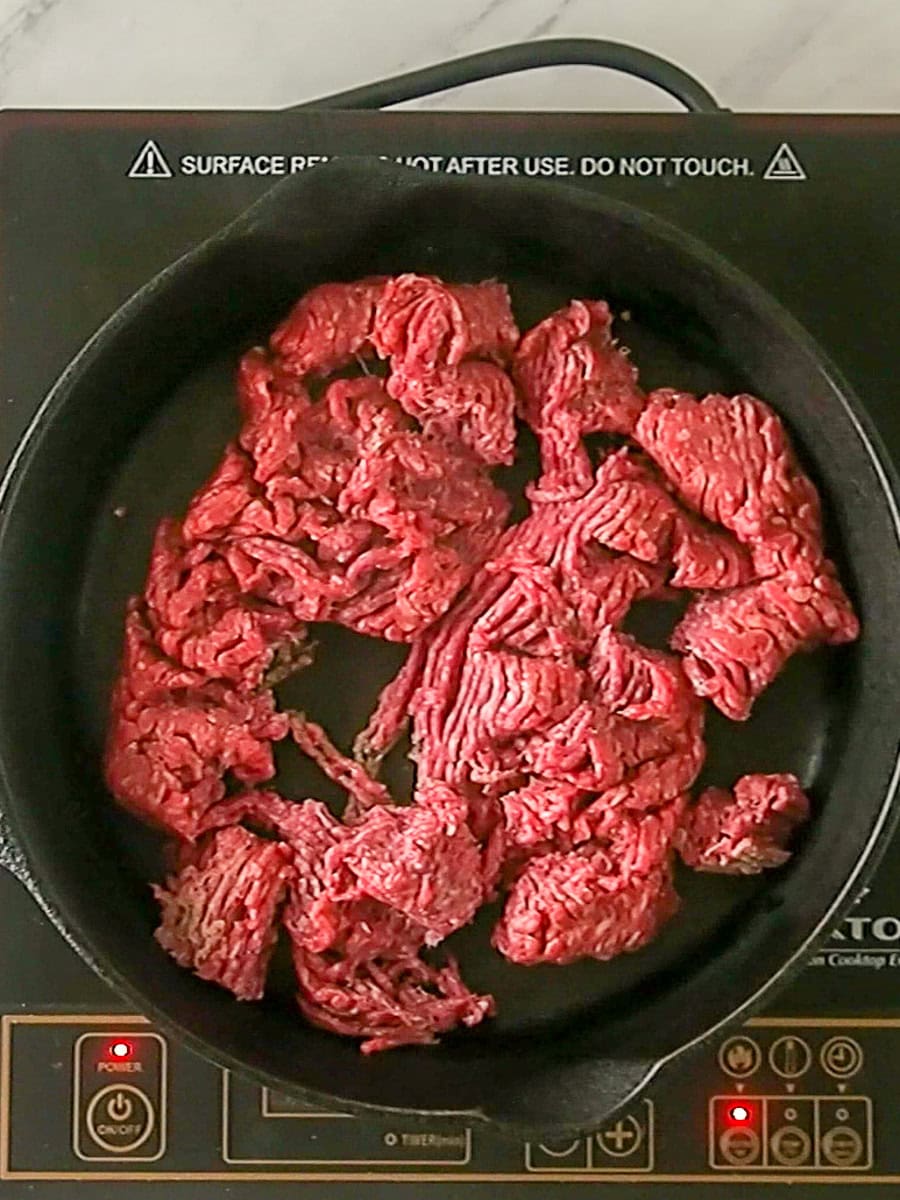 Raw ground beef cooking in a black skillet on an electric stovetop, beginning to brown in some areas.