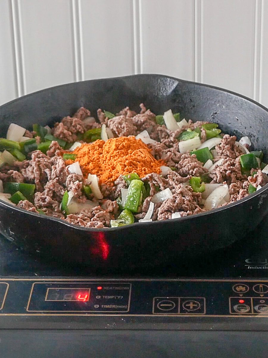 Ground beef, chopped onions, green bell peppers, and taco seasoning are cooking in a black skillet on an induction cooktop, with the temperature set to level 4.