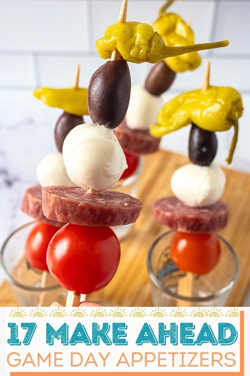 Skewers with pepperoncini, black olive, mozzarella ball, sausage slice, and cherry tomato arranged in glasses on a wooden board.