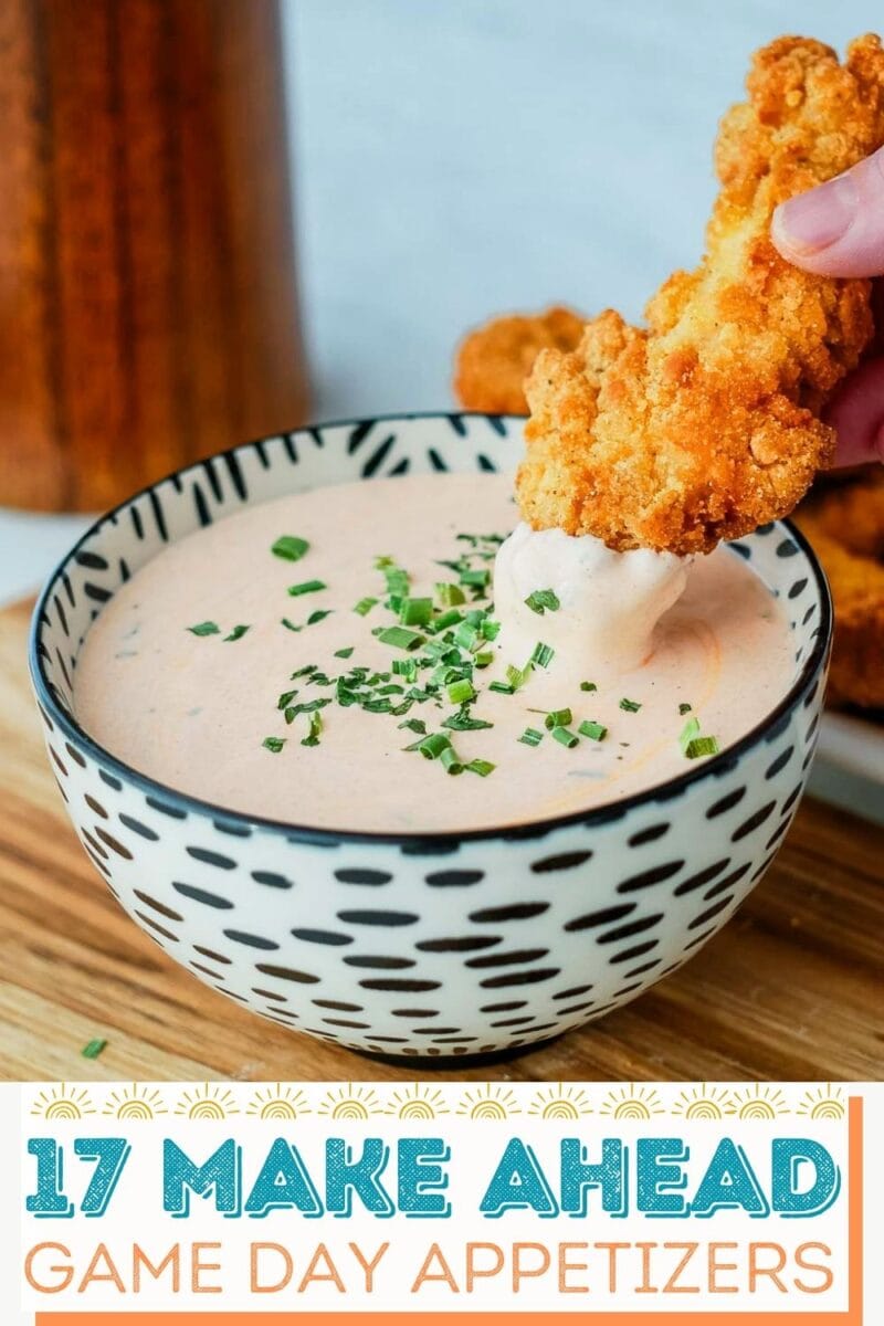 A hand dips a breaded chicken strip into a bowl of creamy dipping sauce, topped with chopped chives. Text below reads, “17 Make Ahead Game Day Appetizers.”.