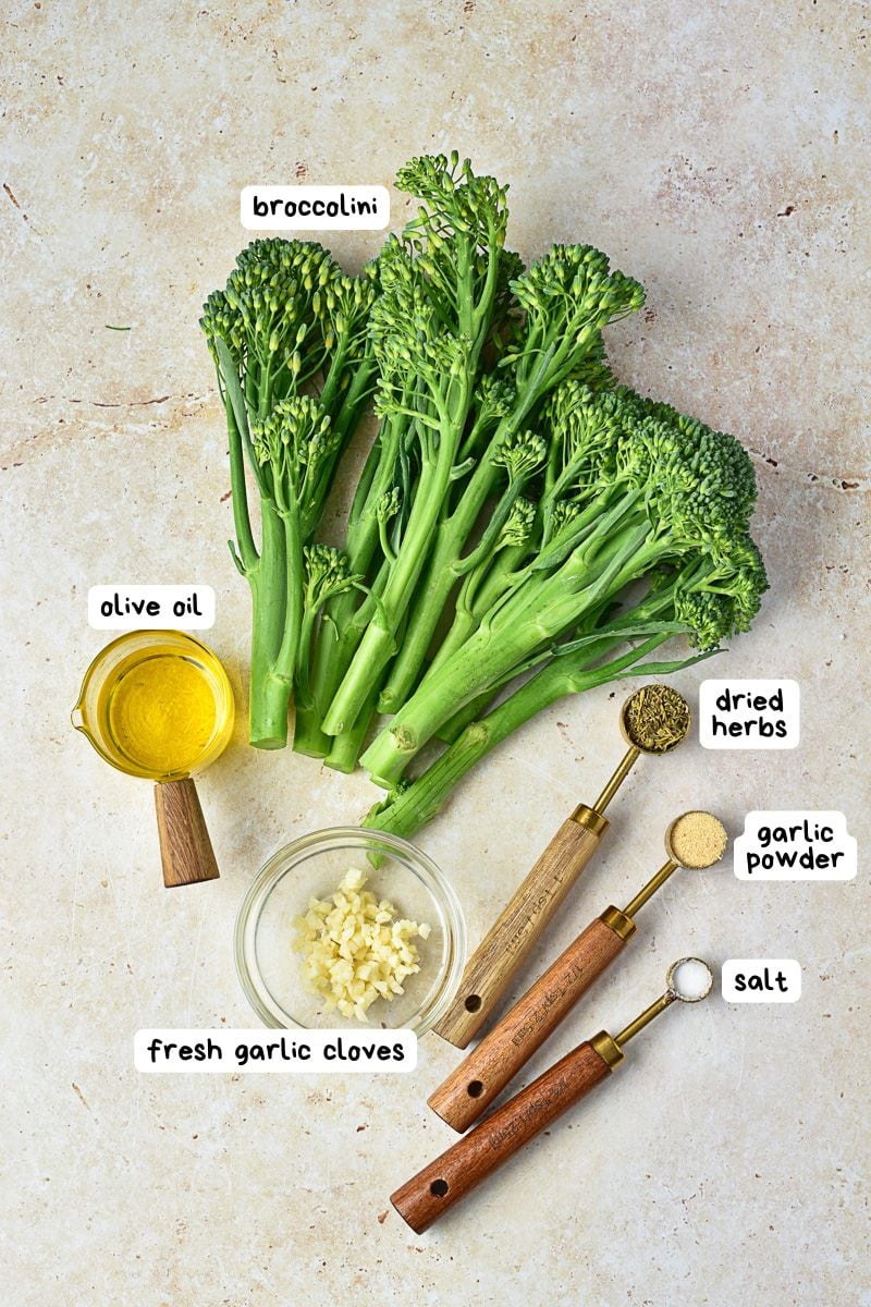 Ingredients for a recipe are arranged on a light surface: broccolini, olive oil, fresh garlic cloves, dried herbs, garlic powder, and salt, each labeled with handwritten-style tags.