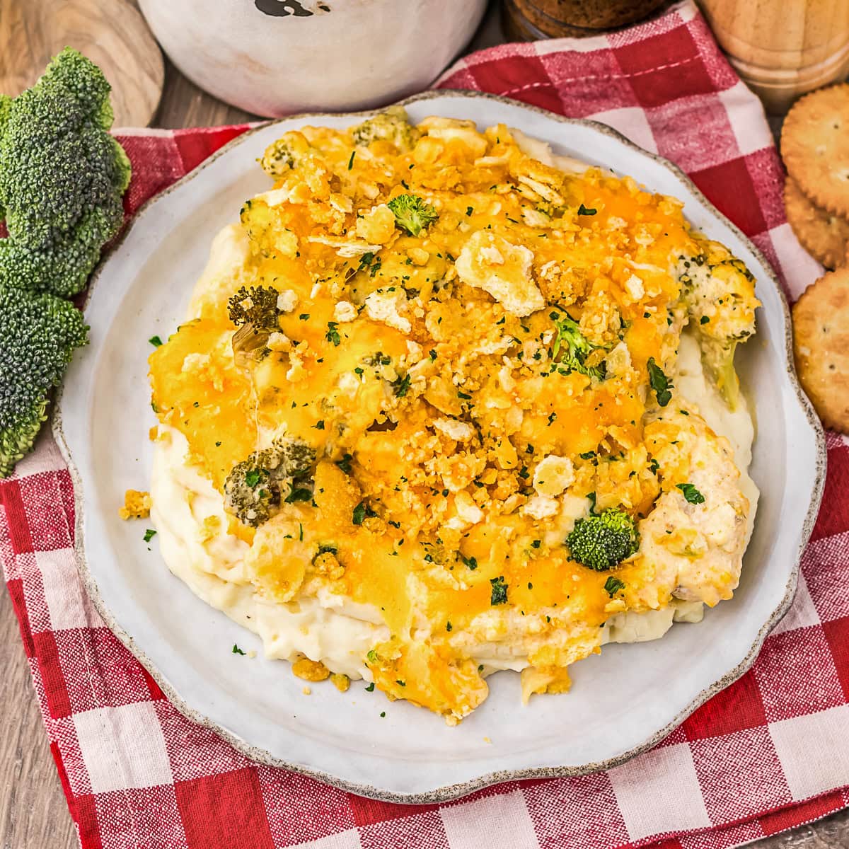 A plate of broccoli casserole with cheese and crushed crackers on top, served on a red and white checkered cloth, with broccoli and crackers nearby.