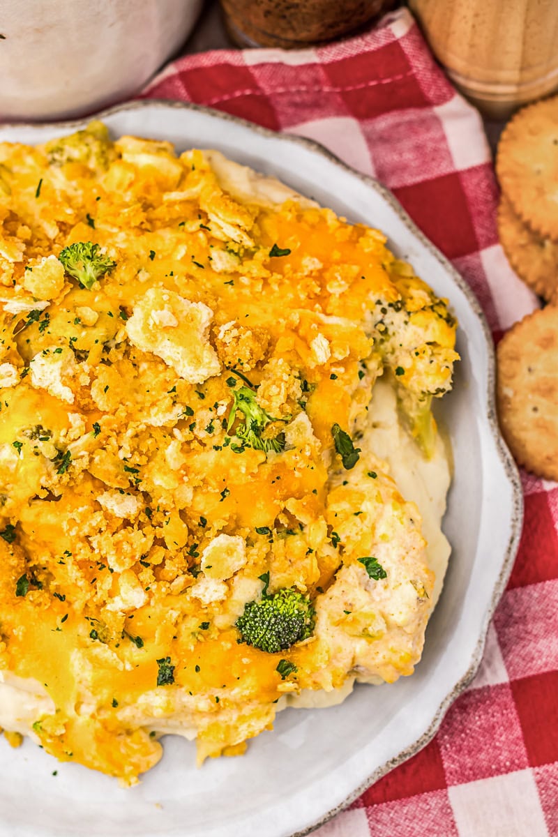 Broccoli and cheese casserole topped with crushed crackers, served on a white plate next to round crackers and a red and white checkered cloth.