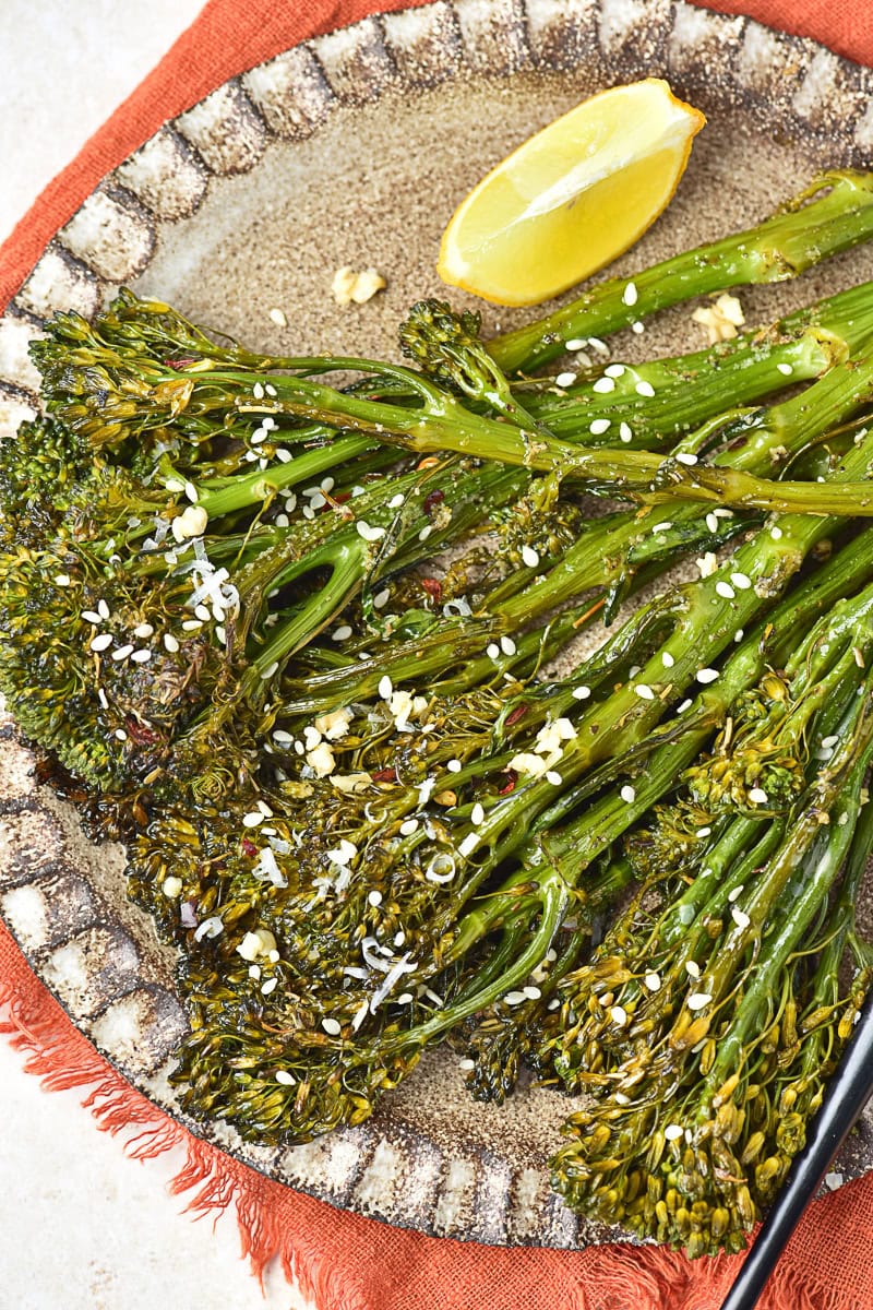 Roasted broccolini garnished with sesame seeds and red pepper flakes on a ceramic plate, served with a lemon wedge.