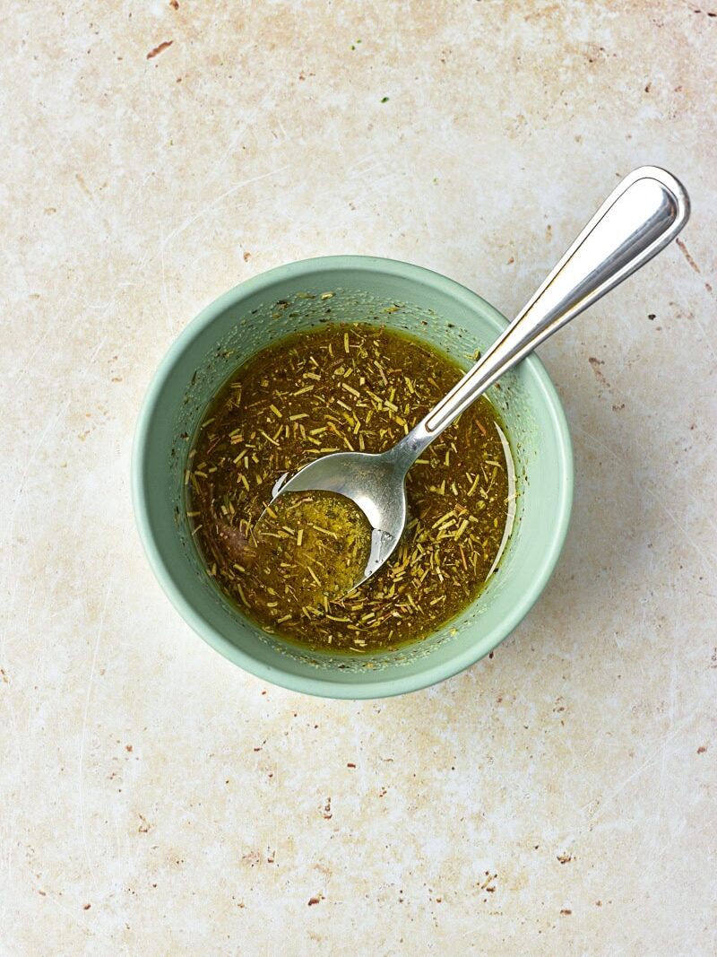 A green bowl with olive oil and herbs mixture, stirred with a spoon, placed on a light-colored textured surface.