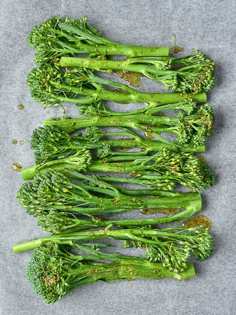 Broccolini stalks arranged on a sheet of parchment paper, drizzled with oil and sprinkled with seasoning, ready to be roasted.