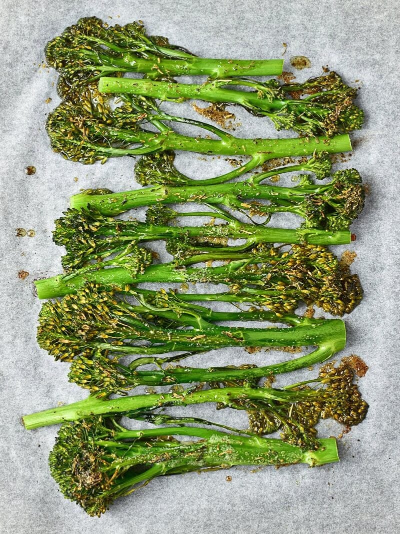 Roasted broccolini arranged in rows on a parchment-lined baking sheet, seasoned with herbs and spices.