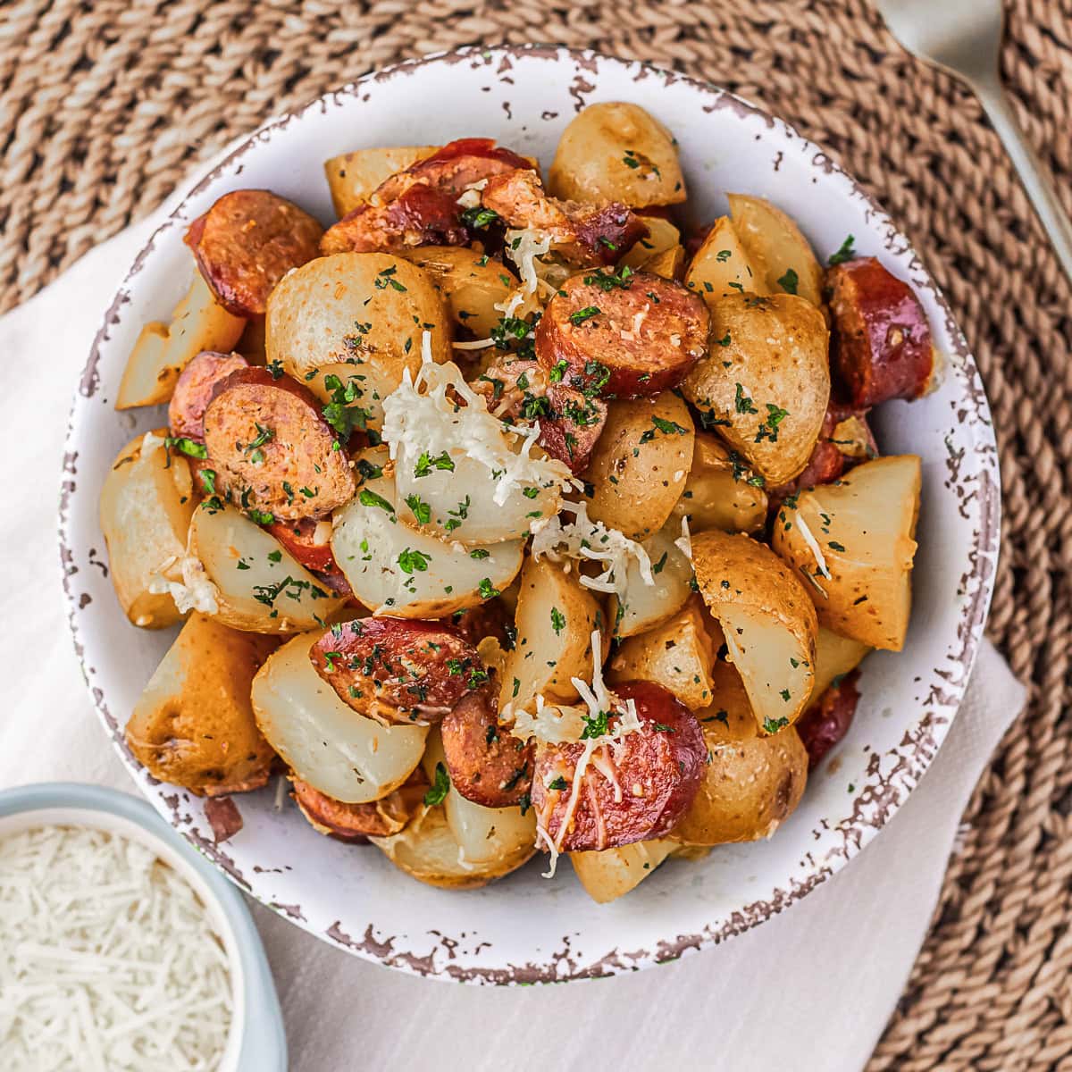 A bowl of roasted potatoes, sausage, and onions garnished with herbs, placed on a woven mat with a small bowl of grated cheese on the side.