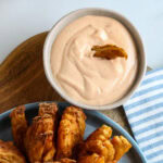 A bowl of creamy dipping sauce with a piece of fried onion dipped in it, next to a plate of fried onion petals on a wooden surface.