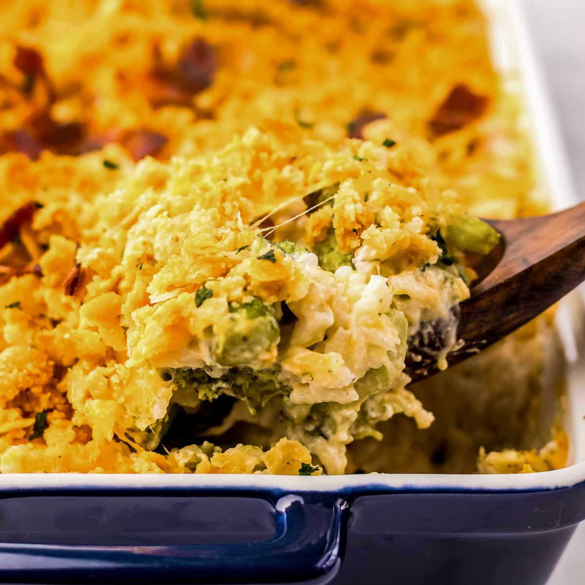A wooden spoon lifts a serving of baked casserole with a golden, crispy topping from a blue dish.