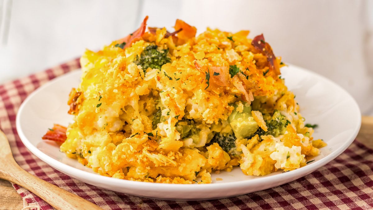 A serving of cheesy broccoli and rice casserole with crispy topping sits on a white plate over a red plaid cloth, next to a wooden spoon.