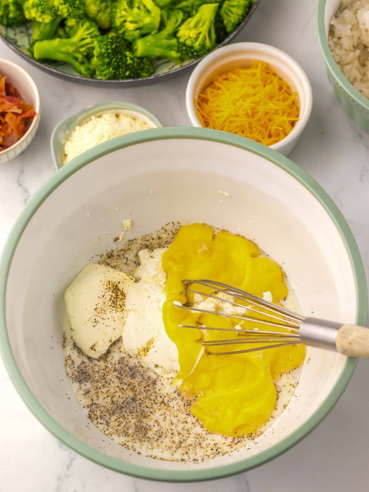A mixing bowl with eggs, cream cheese, sour cream, black pepper, and a whisk. Nearby are bowls of shredded cheese, cooked bacon, broccoli, and rice.