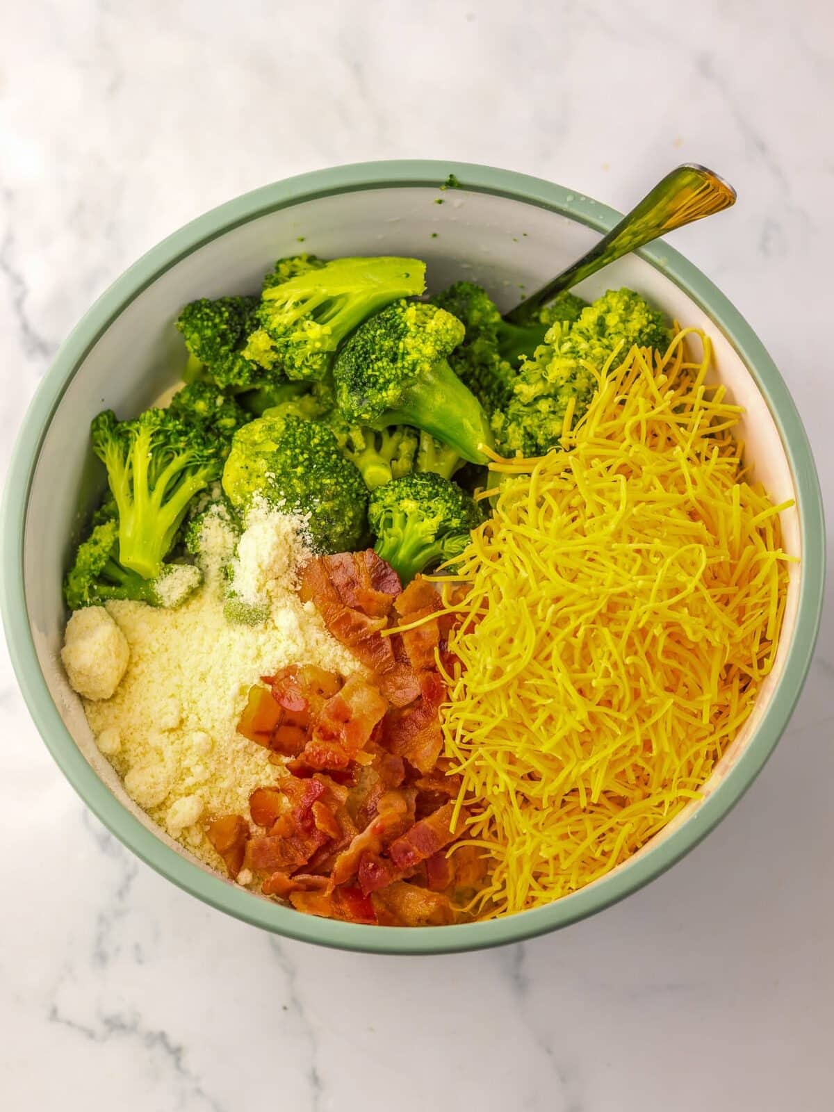 A bowl containing fresh broccoli florets, shredded cheese, crumbled bacon, grated parmesan cheese, and a spoon on a marble surface.