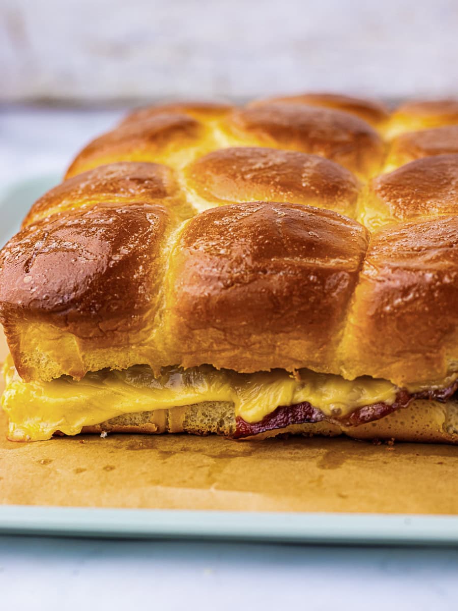 A close-up of a baked cheese and ham slider on a tray, featuring golden brown, shiny bread rolls with melted cheese visible between layers.