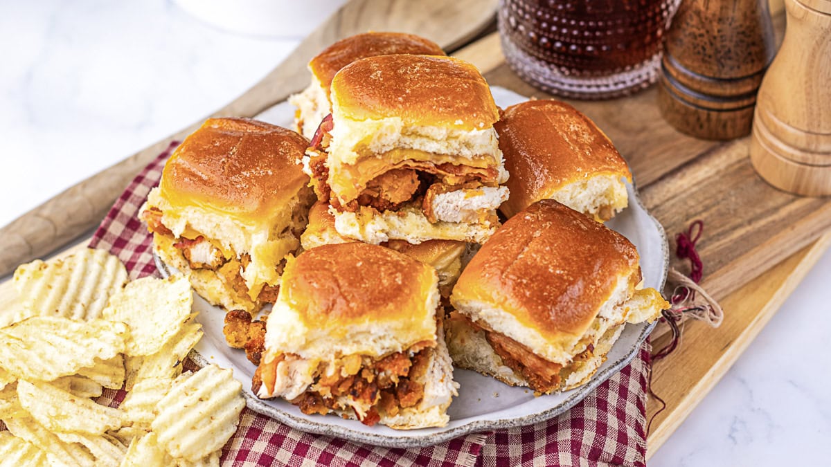 A plate of six chicken sliders with bacon, served on a white plate with a side of ridged potato chips, sits on a red-checked napkin and wooden board.
