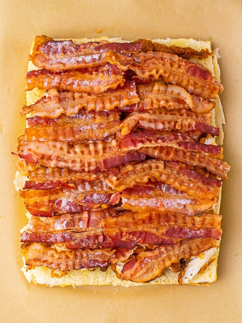 Several strips of cooked bacon are arranged in parallel rows on top of a rectangular piece of bread, all placed on a sheet of brown parchment paper.