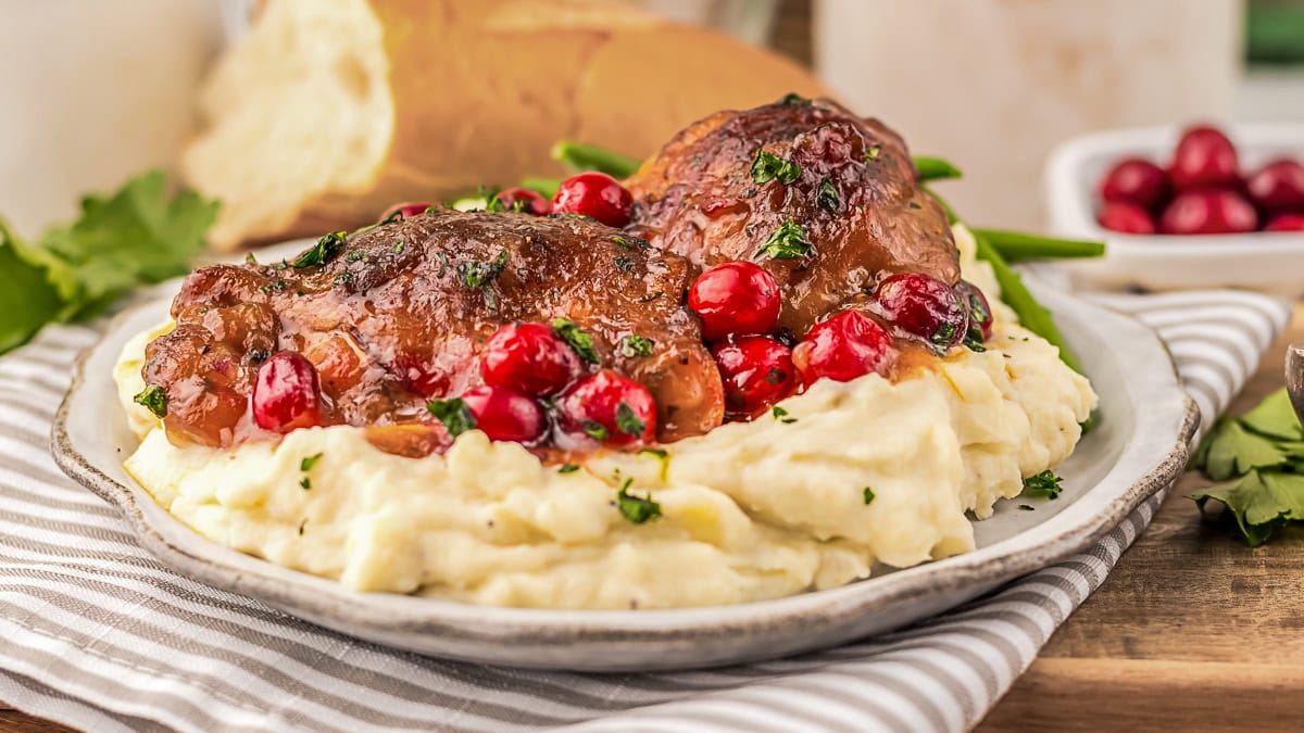 Plate of mashed potatoes topped with braised meat, cranberry sauce, and herbs, with green beans and bread in the background.