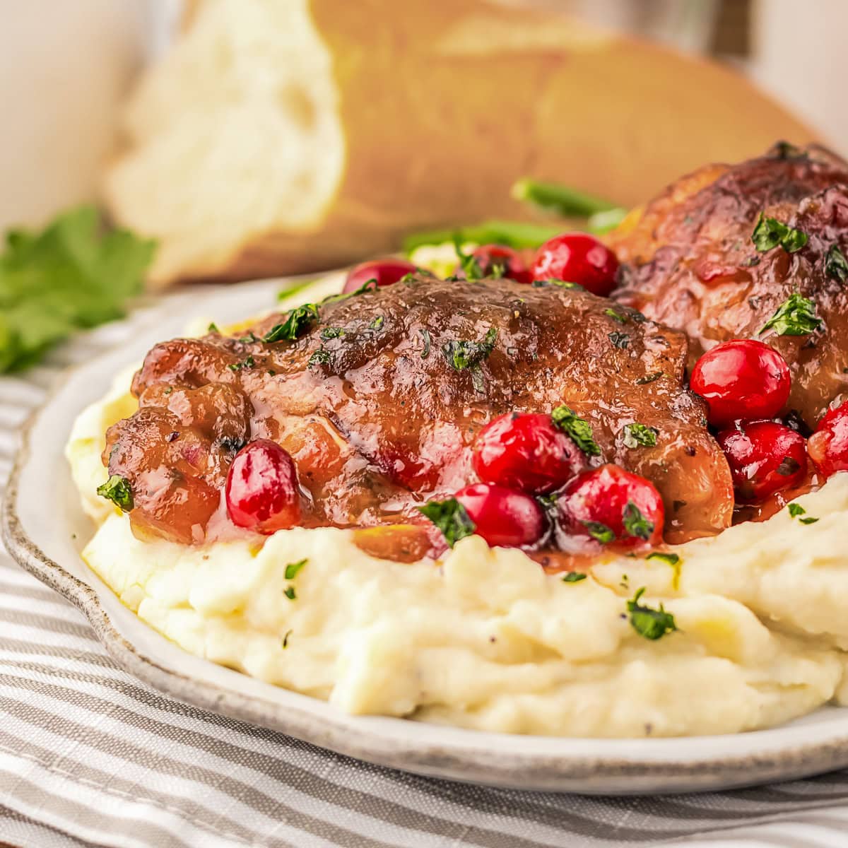 Braised chicken with cranberries served on mashed potatoes, garnished with herbs. A loaf of bread is in the background.