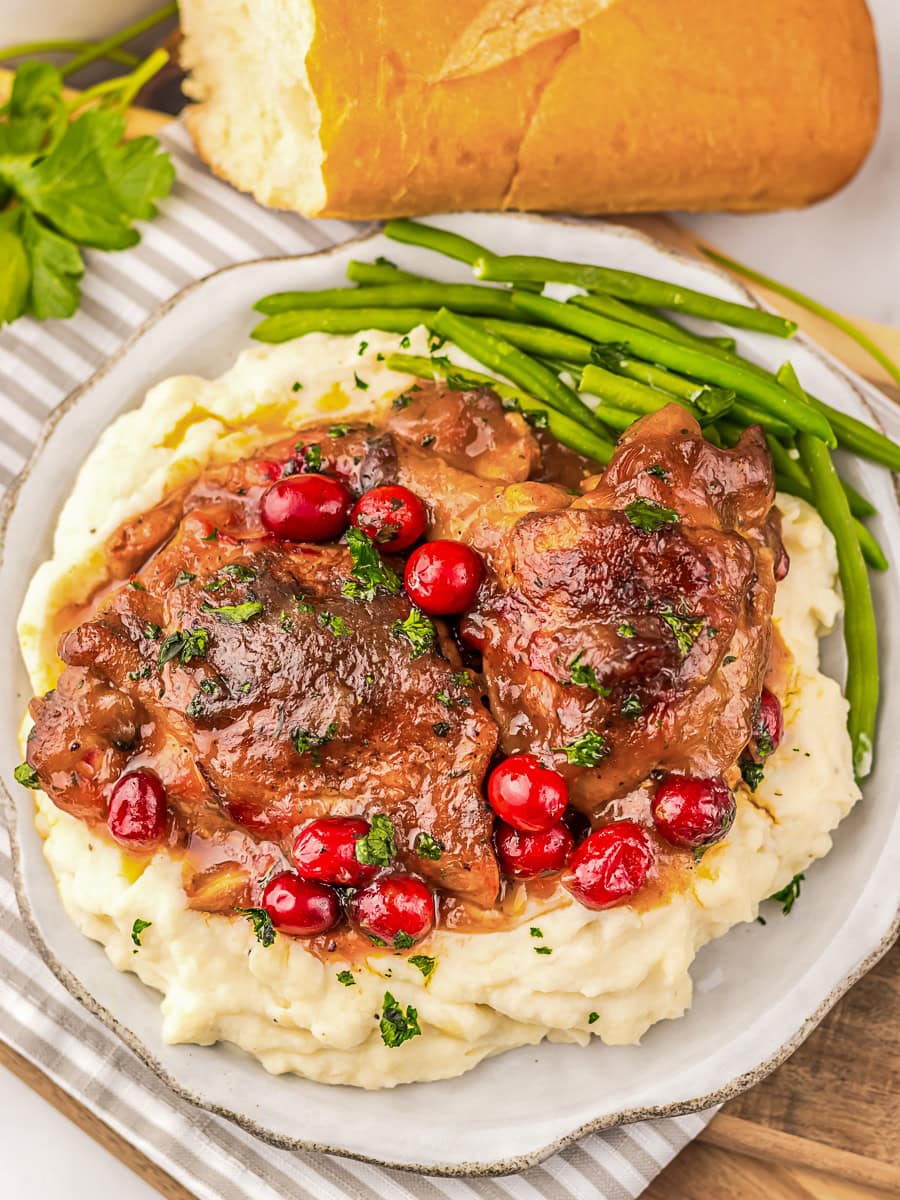 Plate of glazed chicken with cranberries served over mashed potatoes with green beans, garnished with parsley. A loaf of bread and parsley are in the background on a striped towel.