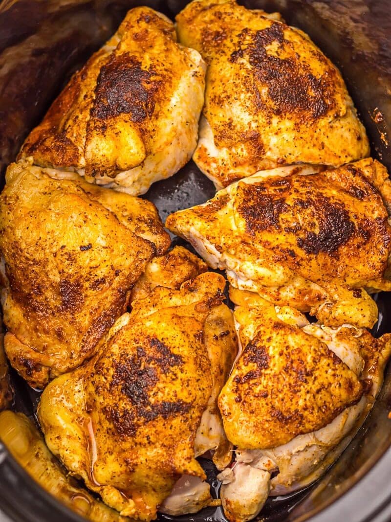 Six seasoned, cooked chicken thighs arranged in a slow cooker with golden-brown, crispy skin and visible cooking juices around the meat.