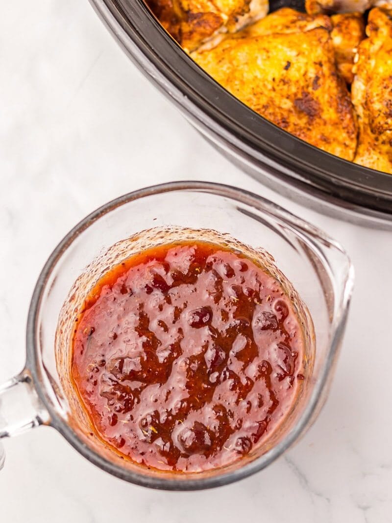 A glass measuring cup filled with a reddish sauce sits on a white surface near a slow cooker containing seasoned chicken.