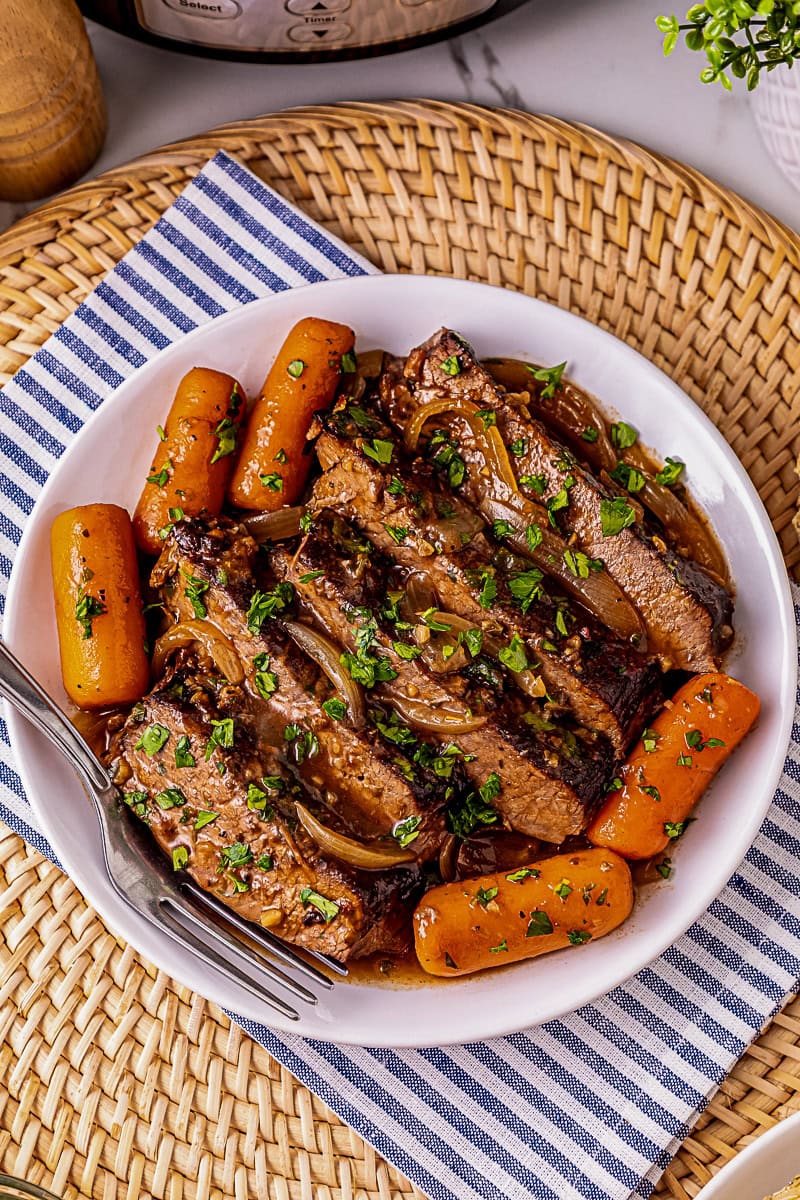 A white plate with sliced beef pot roast, cooked baby carrots, and onion, garnished with chopped parsley, on a woven placemat with a striped napkin and a fork.