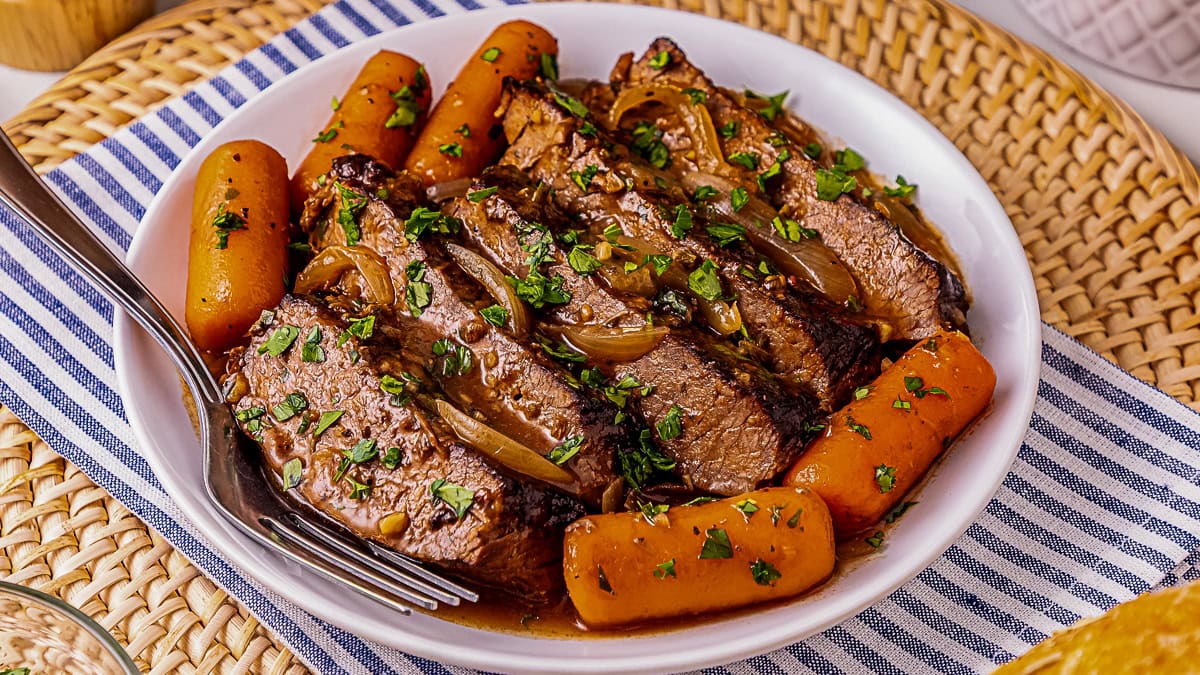 Slices of pot roast with carrots and onions in gravy, garnished with chopped parsley, served in a white bowl with a fork on a striped cloth placemat.