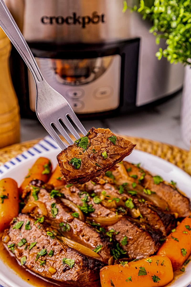 A fork holds a piece of cooked beef above a plate of sliced pot roast with carrots and herbs; a Crockpot slow cooker is in the background.