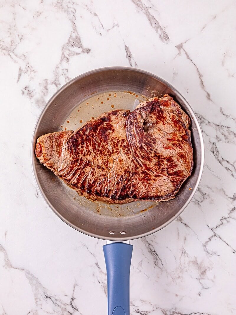 A seared steak is cooking in a stainless steel frying pan with a blue handle, placed on a white marble countertop.