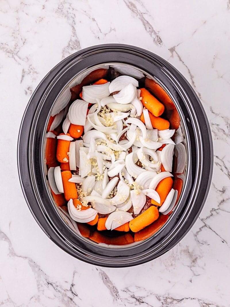 A slow cooker filled with chopped onions, carrots, and minced garlic on a white marble surface.