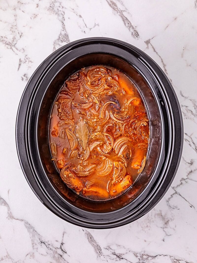 Slow cooker filled with shredded meat, onions, bay leaves, and red-brown broth sits on a white marble surface.