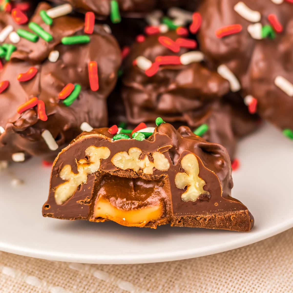 Chocolate candy filled with pecans and caramel, topped with red, white, and green sprinkles, sits on a plate with more candies in the background.